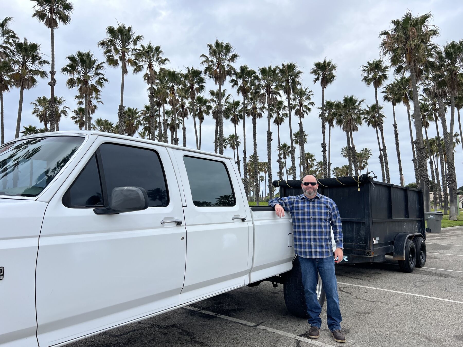 Man standing beside the white Weaver Hauling pickup and black dump trailer in front of a row of palm trees